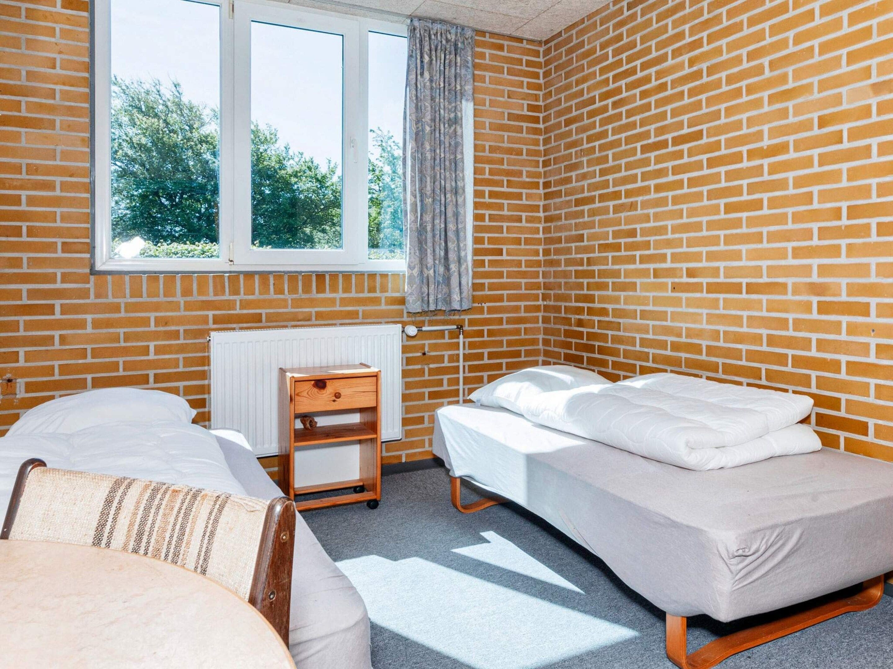 A room with two beds, a window with curtains, a radiator, and a small wooden nightstand.