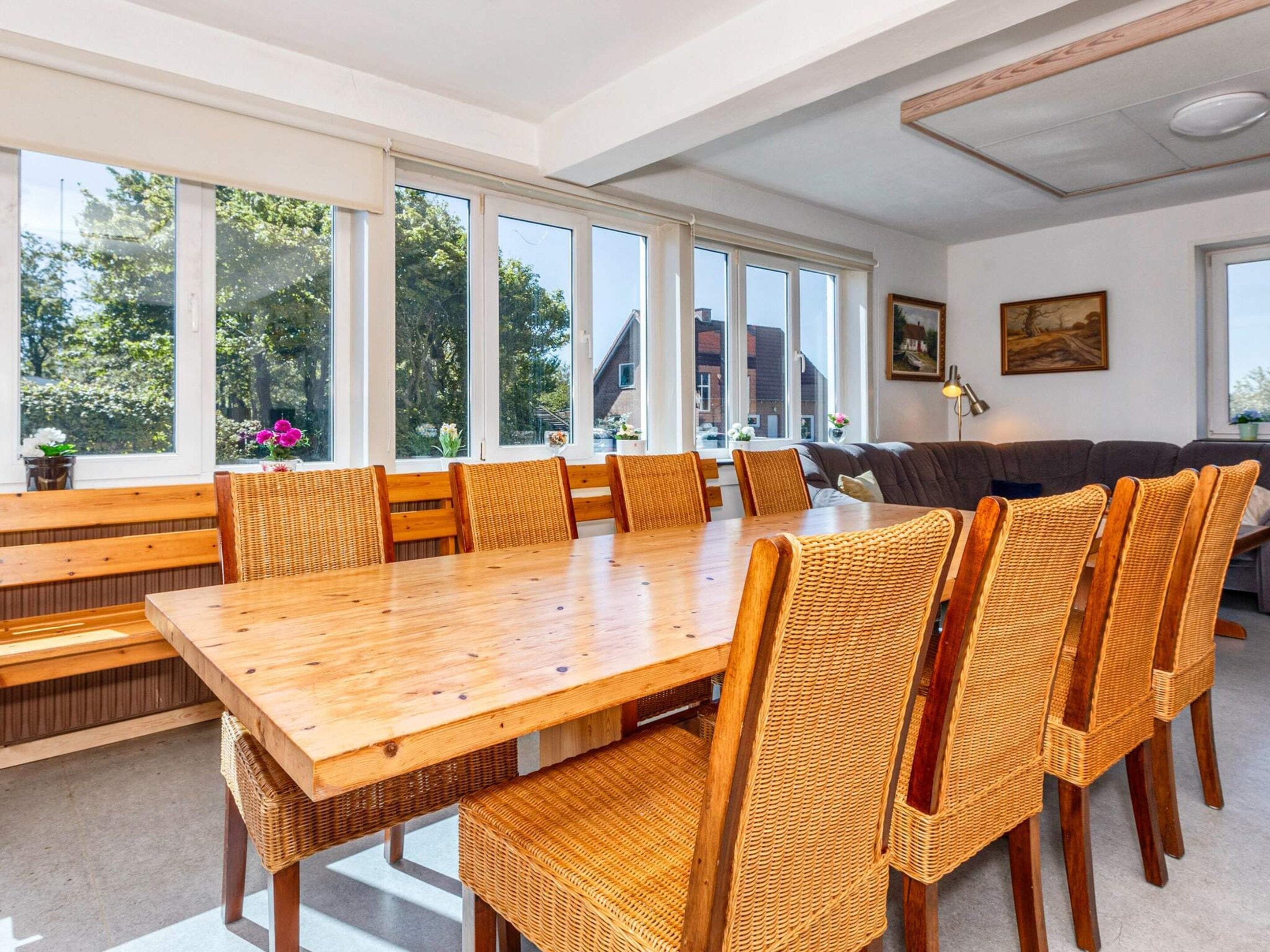A dining area with a large wooden table and wicker chairs, large windows with a view of trees, and a sofa in the background.