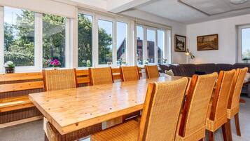 A dining area with a large wooden table and wicker chairs, large windows with a view of trees, and a sofa in the background.