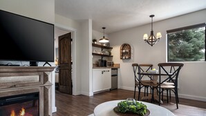 Een woonkamer met een open haard, een televisie, een eethoek met stoelen en een tafel, en een keuken met planken en een raam.
