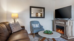 Een woonkamer met een bruine leren bank, een blauwe fauteuil, een witte salontafel met een plant, een open haard en een flatscreen televisie.