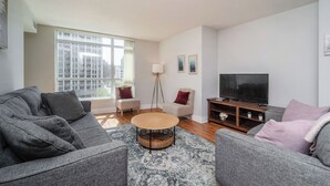 A modern living room with a grey sofa, a round coffee table, a TV, and a large window offering a city view. - Spacious 3bed/2 bath Downtown Toronto condo + Free Parking, min to CN tower (Toronto)