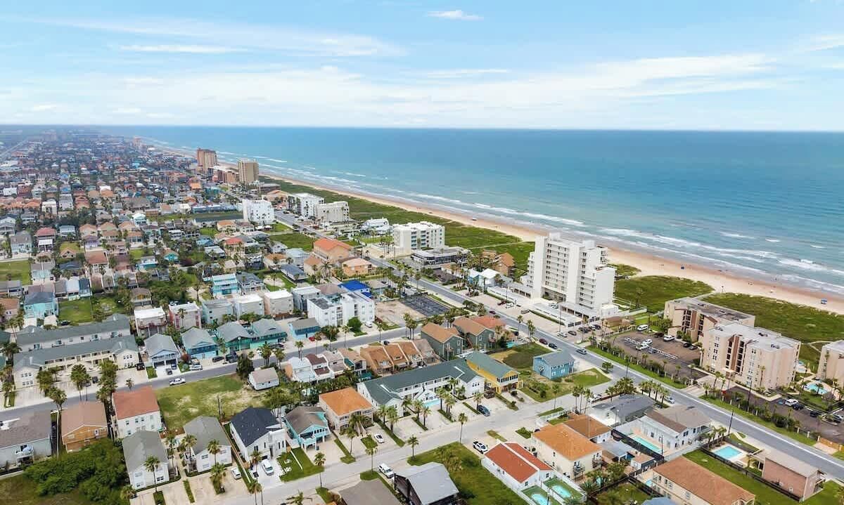A coastal residential area with a beachfront, featuring a mix of single-family homes and multi-story buildings, with a clear view of the ocean.