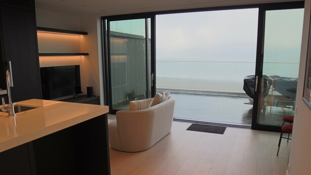 A modern kitchen with a large island, a flat-screen TV, and a sliding glass door leading to a balcony with a view of the sea.
