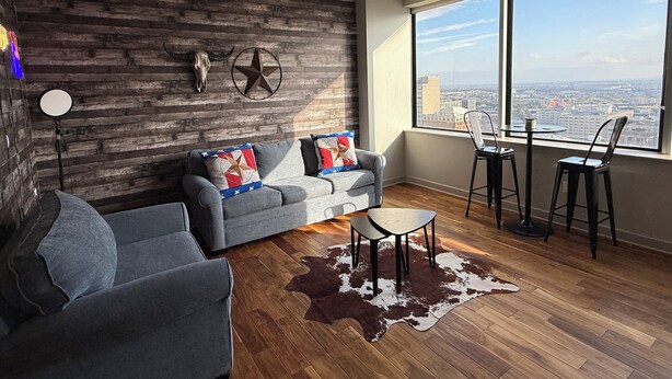 A modern living room with a grey sofa, a coffee table, and a cowhide rug. There is a large window with a city view. - Family-friendly Dallas getaway with rooftop pool, free parking & Texas flair. (Dallas)