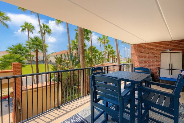 Un balcon avec une table et des chaises, un mur en briques et une vue sur des palmiers et une pelouse verdoyante.