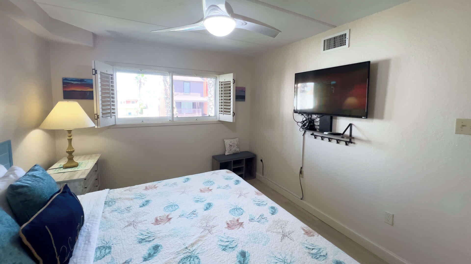 A bedroom with a bed, a nightstand with a lamp, a TV mounted on the wall, and a window with shutters.