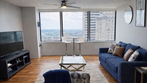 A modern living room with a TV, sofa, and a view of a cityscape through large windows. - Cozy Retreat with Rooftop Pool, Parking, and Spacious Comfort (Dallas)