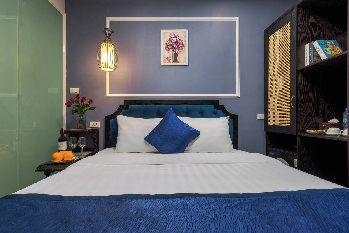 A neatly made bed with a blue and white color scheme, a framed floral artwork on the wall, and a bookshelf with books and decorative items.