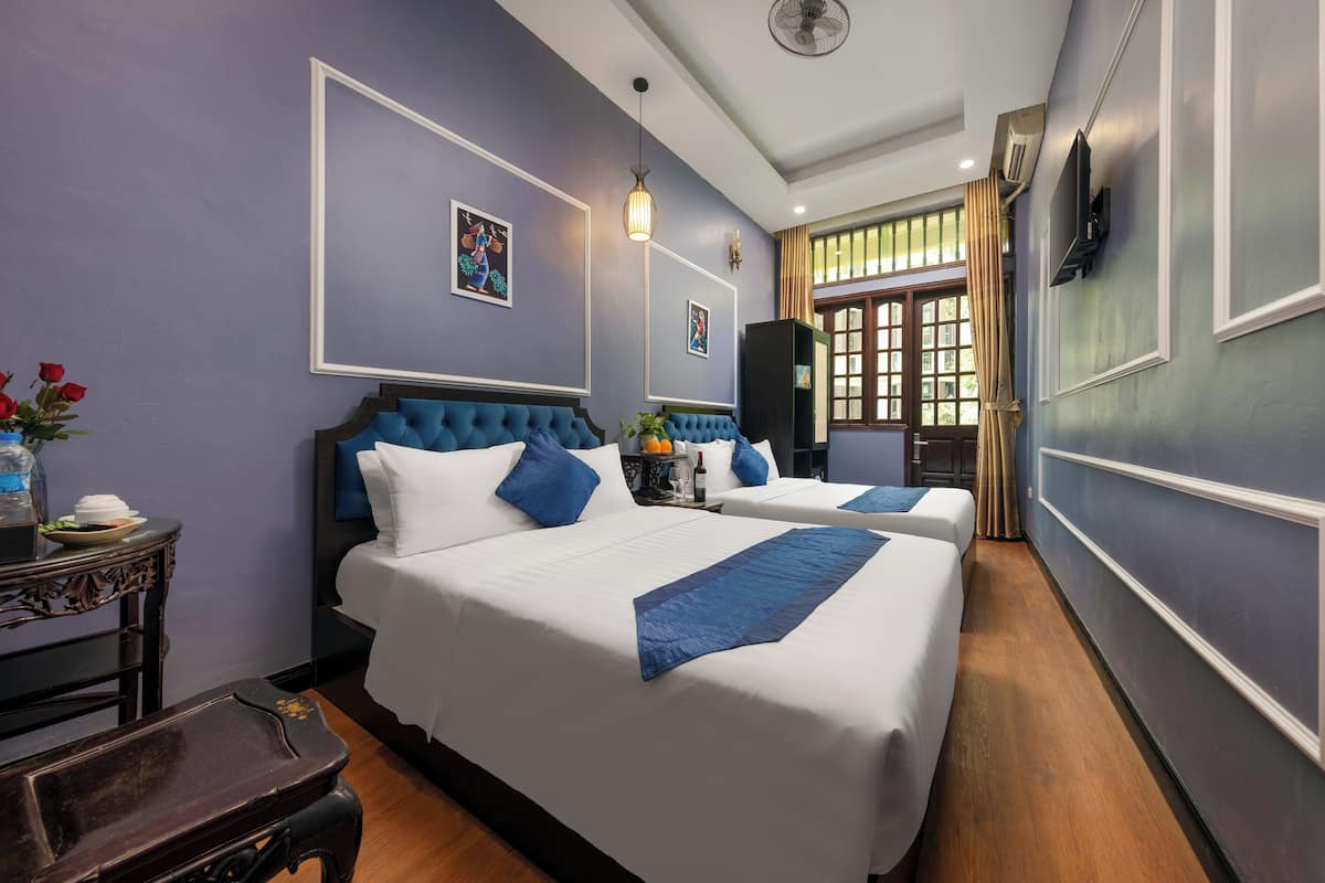 A hotel room with two beds, a large window with curtains, a wall-mounted TV, and a small table with a vase of flowers.