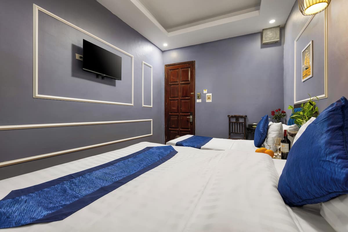 A hotel room with two beds, a TV mounted on the wall, a wooden door, and a small table with a bottle and glasses.