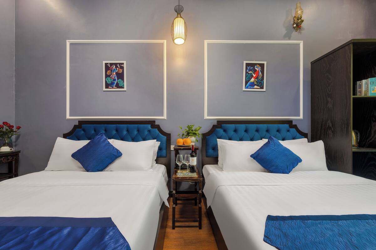 A hotel room with two beds, a small table with a bottle and glasses, and two framed pictures on the wall.