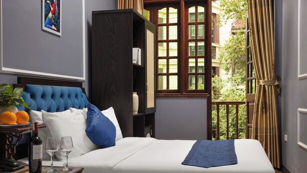 Een hotelkamer met een groot bed, een nachtkastje met een wijnfles en glazen, een boekenkast en uitzicht op groen.