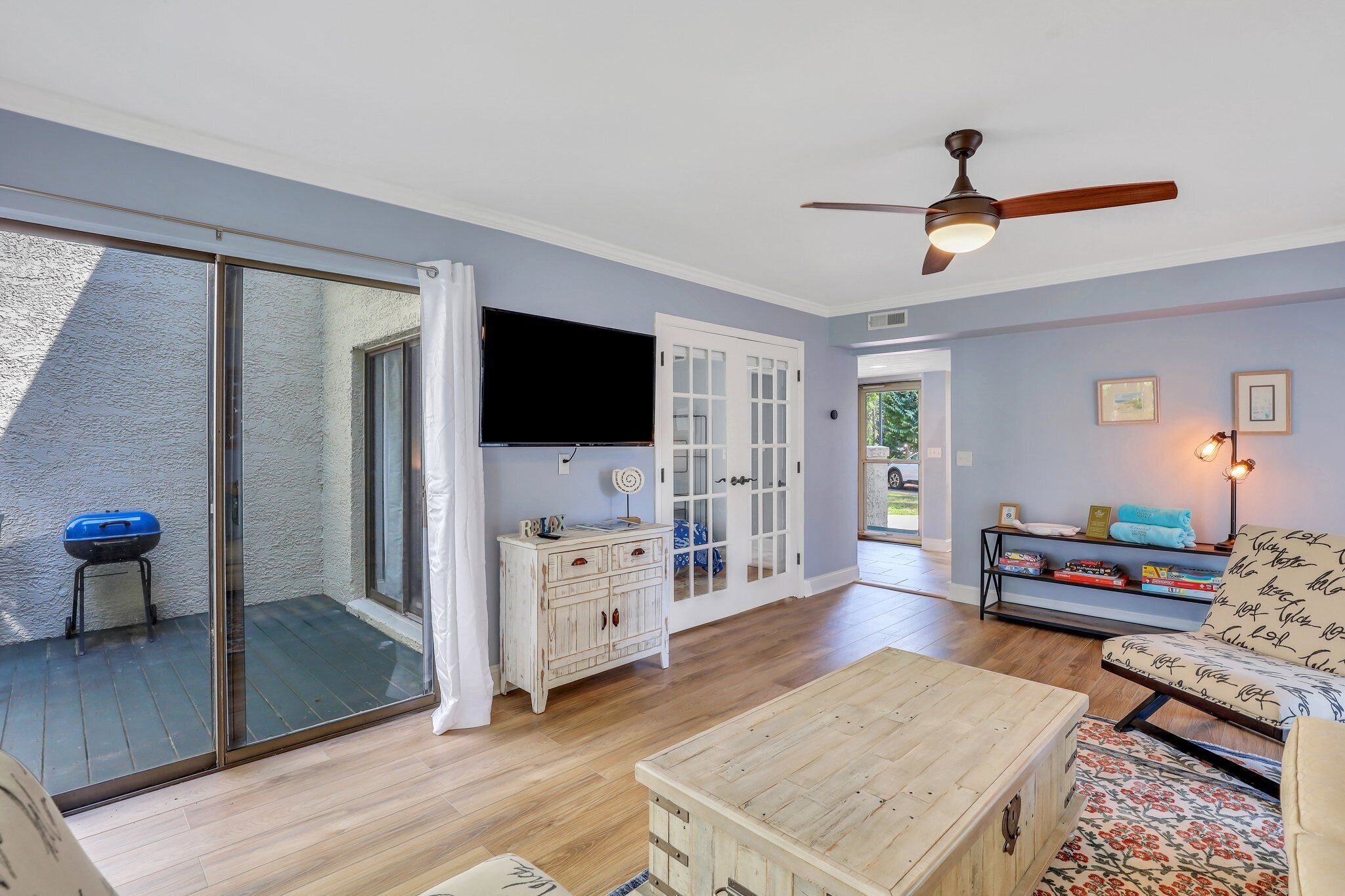 A living room with a sliding glass door leading to a balcony, a ceiling fan, a flat-screen TV, a wooden coffee table, and a sofa with patterned cushions.