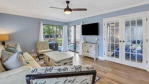 A living room with a sofa, armchair, coffee table, and a TV. There is a ceiling fan, a cabinet, and a sliding glass door leading to an outdoor area. - Luxury Hilton Head Townhome with pool and biking distance to beach (Hilton Head Island)