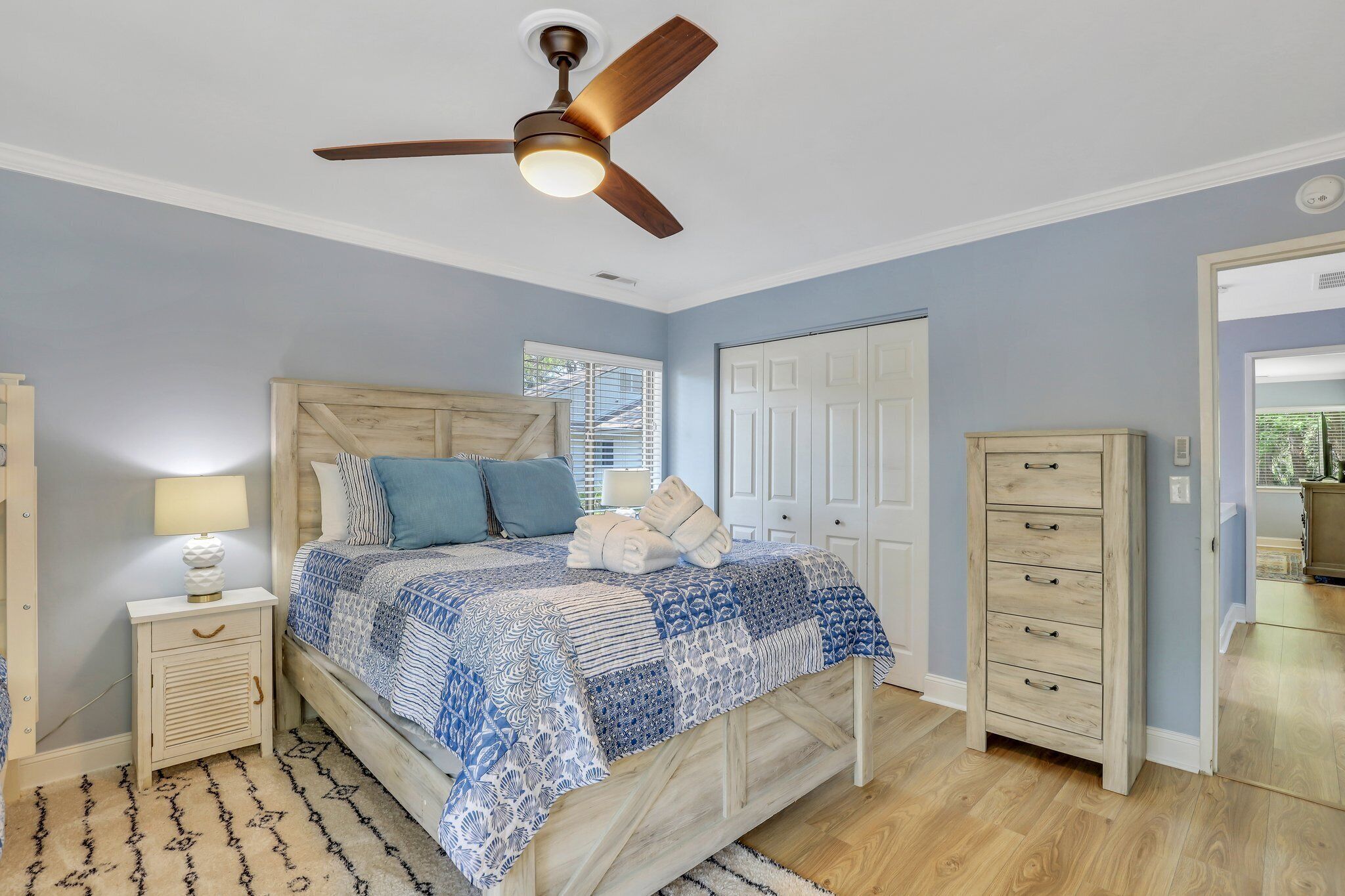 Une chambre à coucher comprenant un lit, une table de chevet, une commode, un ventilateur de plafond et une porte.