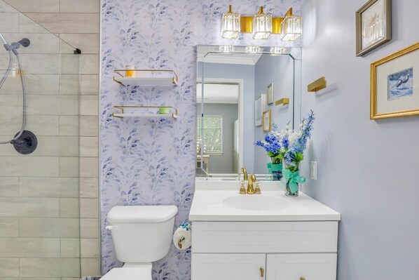 Un baño con inodoro, lavabo con detalles dorados y un espejo sobre él. Hay dos estantes con marcos dorados en la pared, y un florero con flores azules sobre la encimera del lavabo.