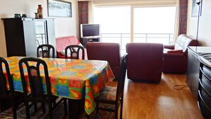 A dining area with a table covered in a colorful tablecloth, surrounded by chairs. There is a sofa and a television in the background. - Apartment in Nieuwpoort Near the Beach (Nieuwpoort)