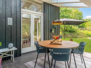 An outdoor patio with a round wooden table, four chairs, a hanging lamp, and a small table with a vase of flowers.