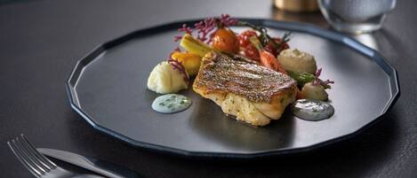 Ein Teller mit Essen, darunter ein Fischstück, Gemüse und Soße, auf einem dunklen Tisch, daneben liegen Gabel und Messer.