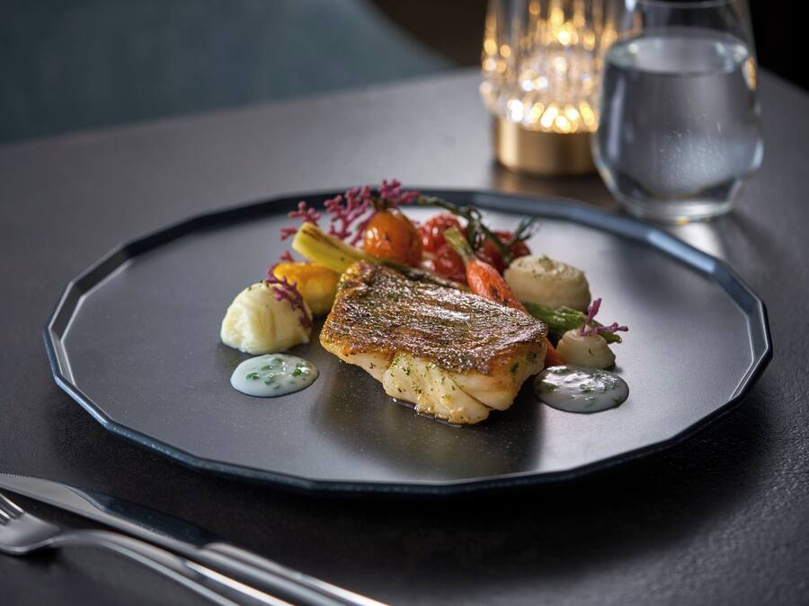 A plate of food with a piece of fish, vegetables, and a sauce, on a dark table with a fork and knife beside it.