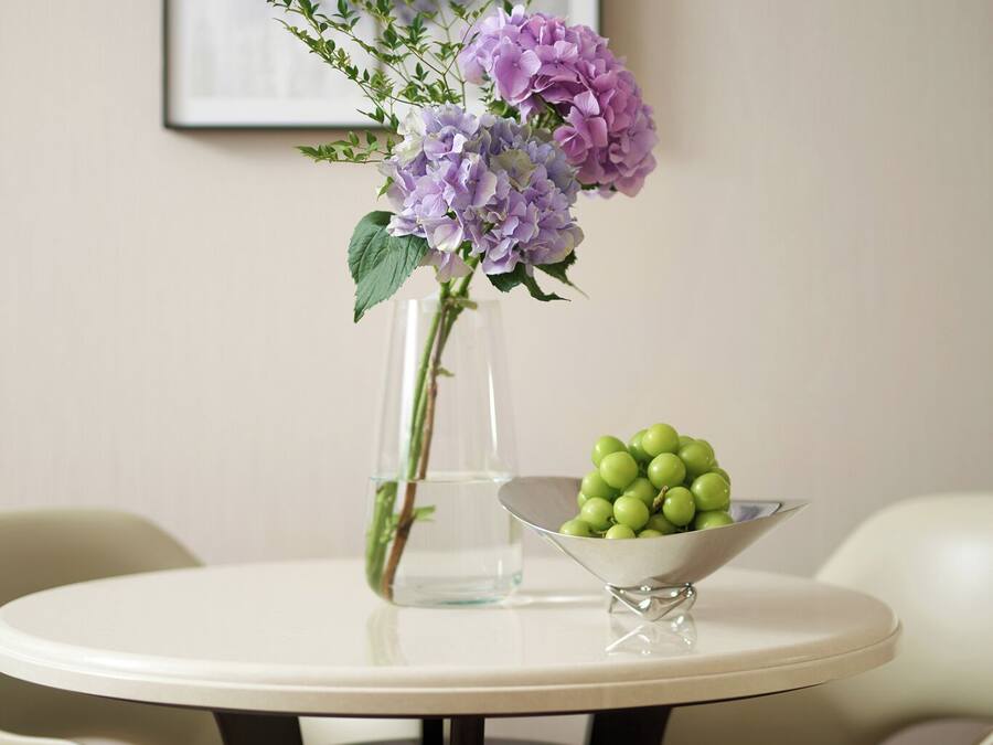 Una mesa redonda con un florero transparente que contiene flores moradas y un bol de uvas verdes.