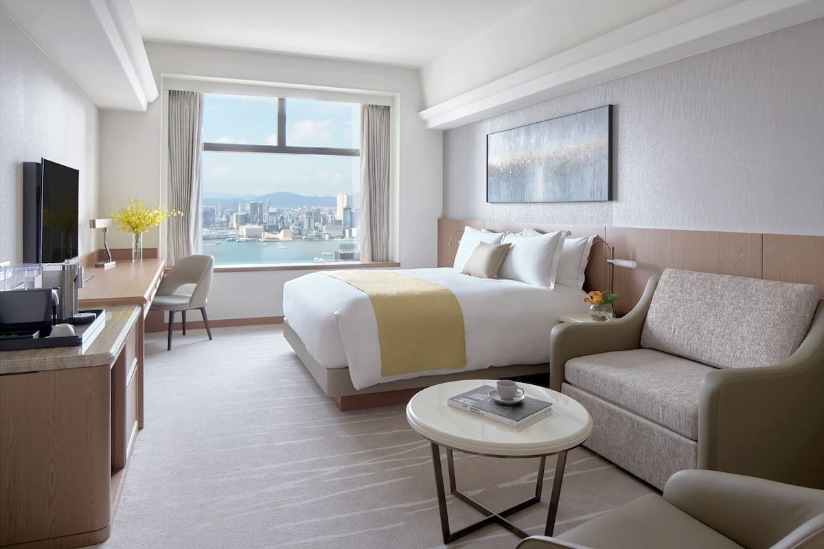 A hotel room with a bed, a desk, a chair, a sofa, and a view of a cityscape through a large window.