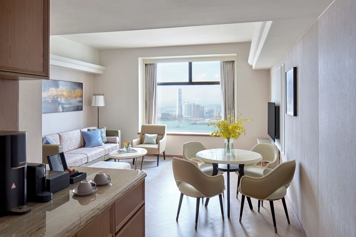 A modern hotel room with a sofa, dining table, chairs, a coffee machine, and a city view.