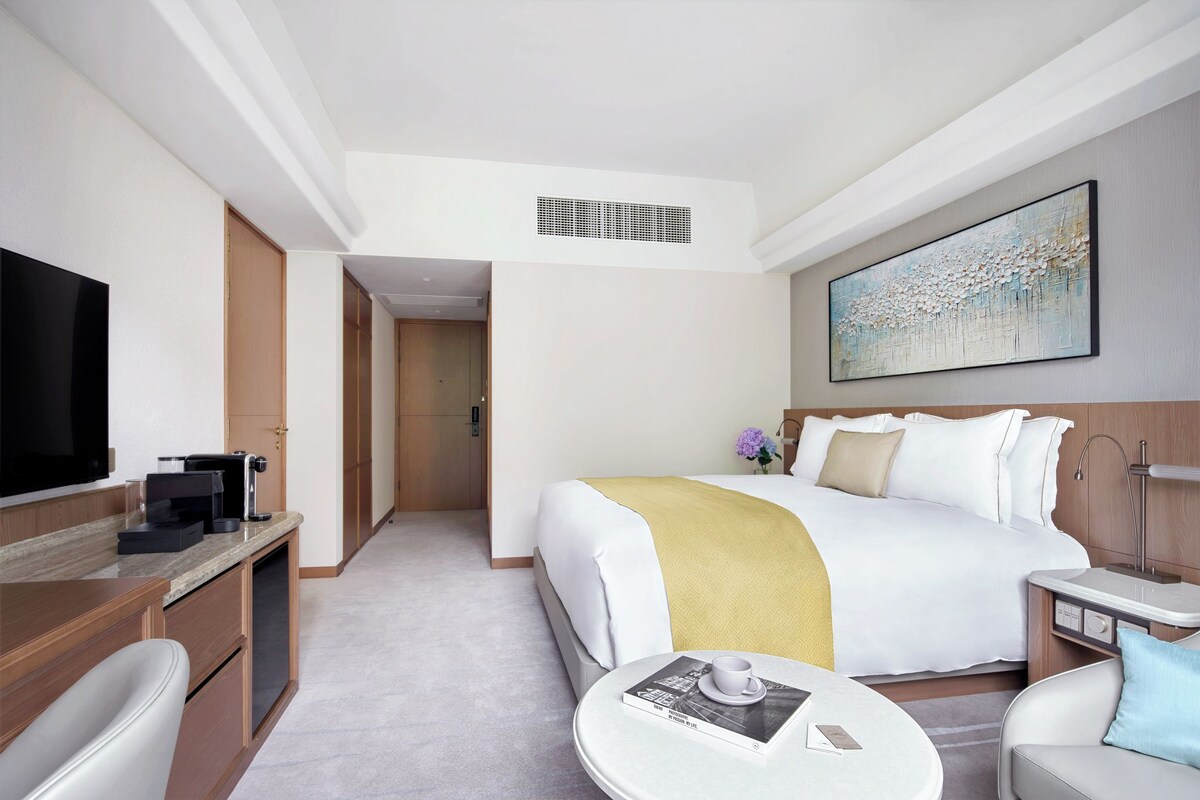 A hotel room with a bed, a TV, a coffee machine, a desk, and a bedside table with a cup and a book.