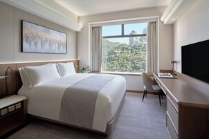 A hotel room with a large bed, a desk, a TV, and a window with a view of a forest and buildings.