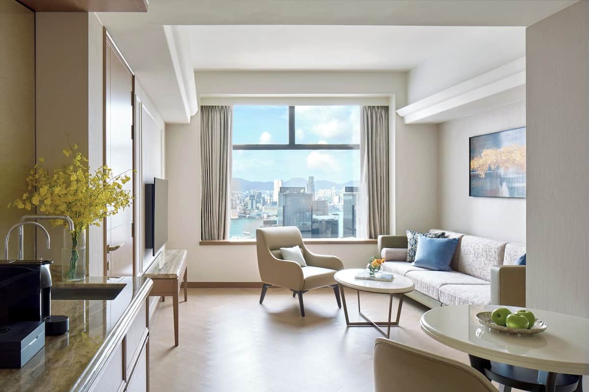 A modern hotel room with a city view, a sofa, a dining table, and a kitchen area.