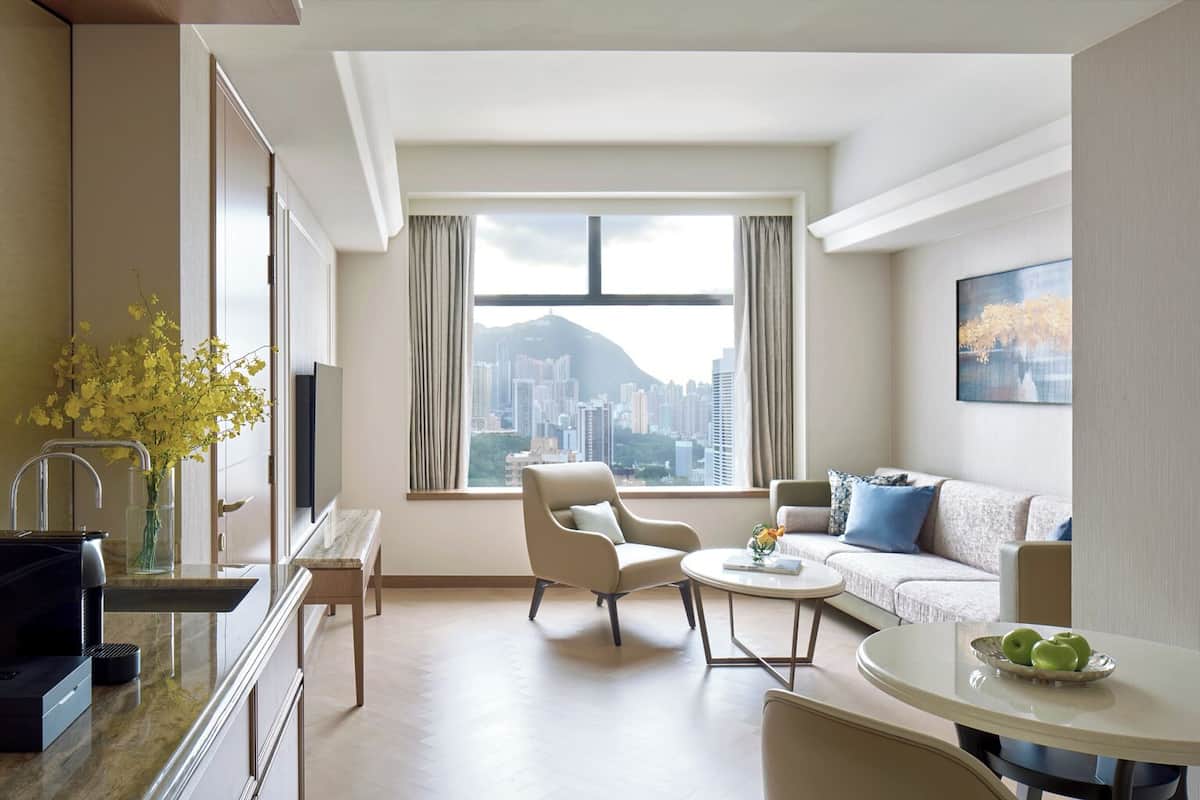 A modern hotel room with a city view, a sofa, a dining table, and a kitchen area.