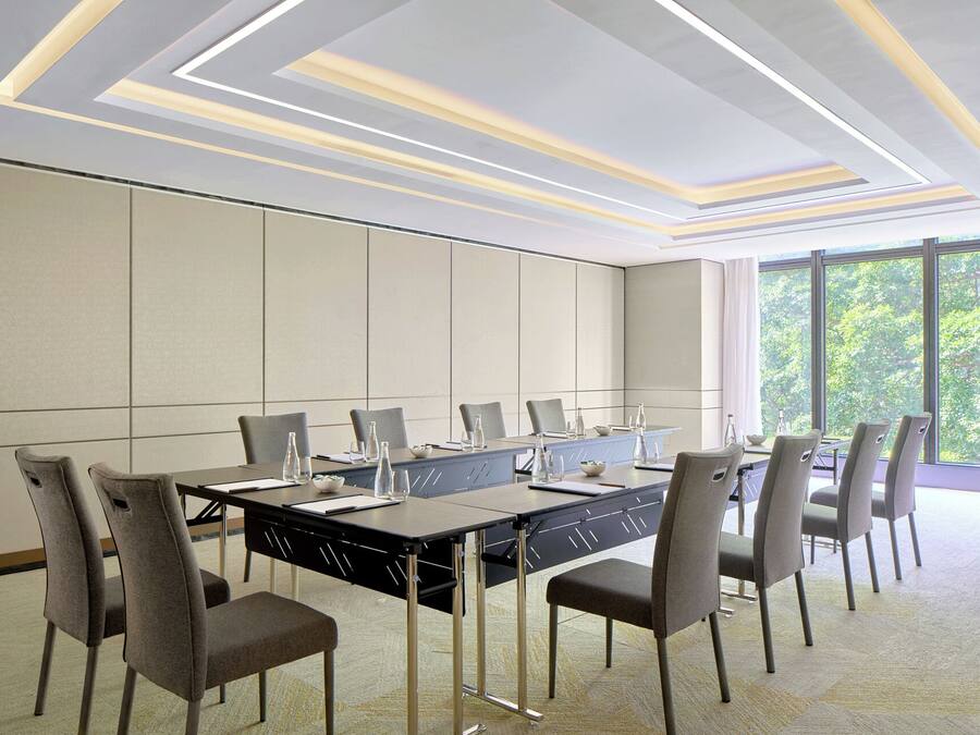A conference room with a long table, grey chairs, and a view of trees through large windows.