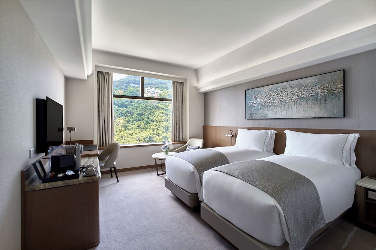 A hotel room with two beds, a desk, a chair, a TV, and a large window with a view of greenery.