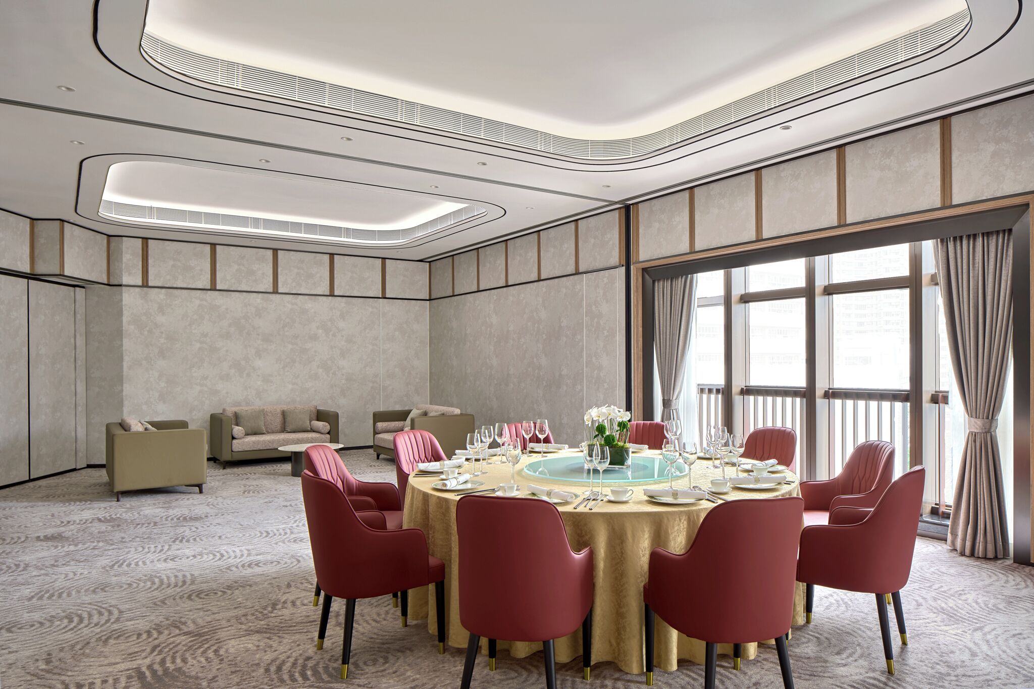 Una camera d'albergo moderna con un tavolo da pranzo rotondo apparecchiato per sei persone, sedie rosse, un divano e un'ampia finestra con tende.
