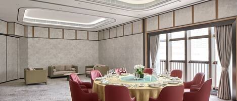 A modern hotel room with a round dining table set for six, red chairs, a sofa, and a large window with curtains.