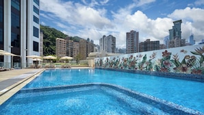 Una piscina con sillas de descanso y sombrillas, rodeada de edificios altos y un mural tropical.