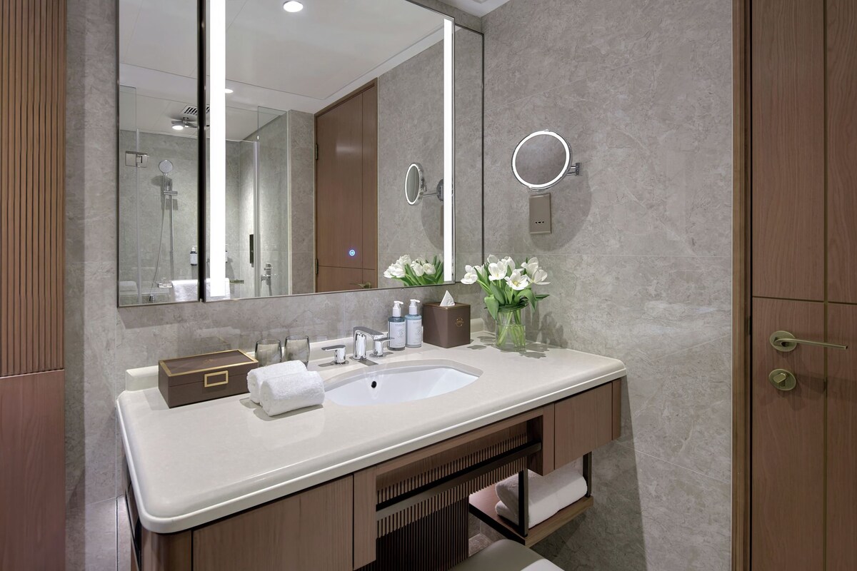 A modern bathroom with a large mirror, a sink, and a vanity. There are toiletries on the counter, a towel, and a vase with flowers.
