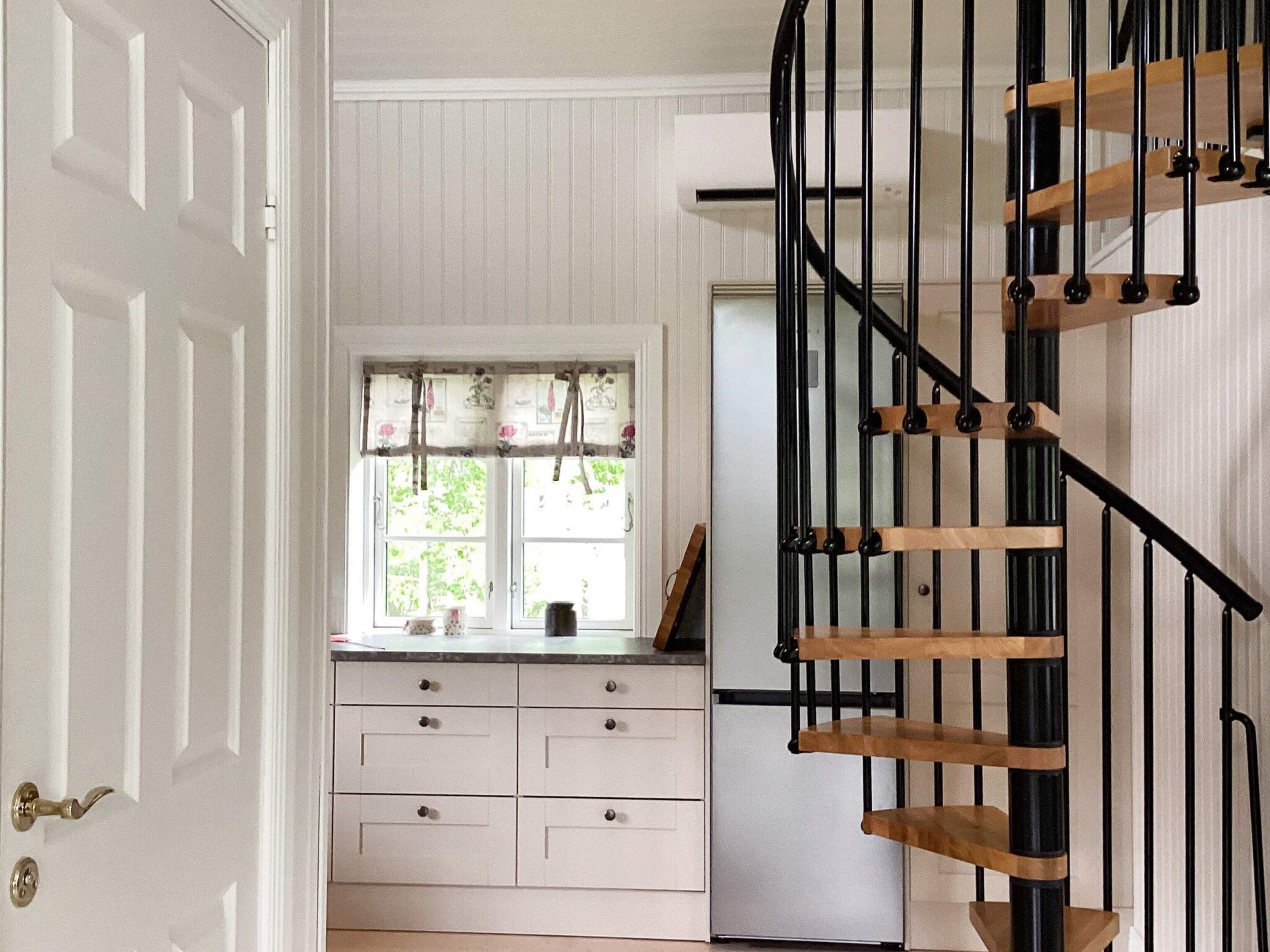 A kitchen with white cabinetry, a window with floral curtains, and a black spiral staircase.