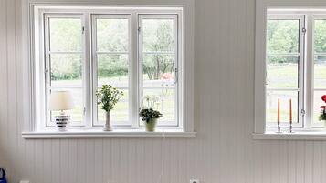 A room with two windows, each with a white frame and a view of greenery outside. There are two potted plants on the windowsill, and a blue bag is visible on the floor.