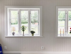 A room with two windows, each with a white frame and a view of greenery outside. There are two potted plants on the windowsill, and a blue bag is visible on the floor.