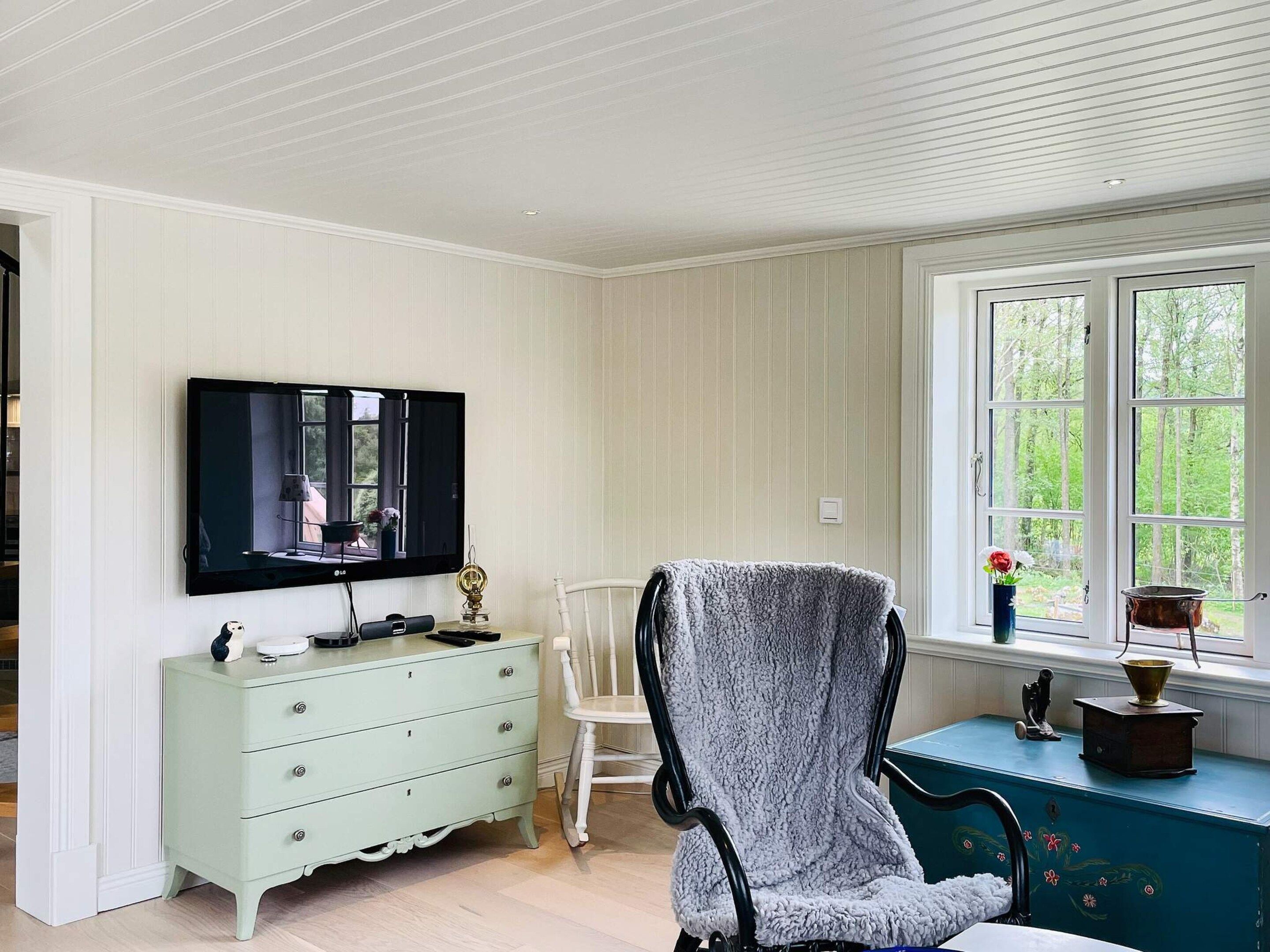 A room with a television on the wall, a white chair, a light green dresser, and a window with a view of greenery.