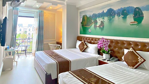 A hotel room with two beds, a balcony with a view, a painting of a coastal scene, and a fan. - Blue Diamond Hotel (Hong Gai)