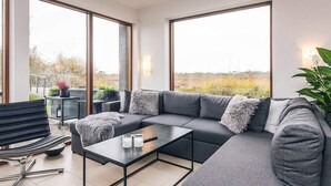 A modern living room with a grey sectional sofa, a glass coffee table, and a black metal-framed table. Large windows offer a view of the outdoors. - 4 Star Holiday Home in Bogense (Bogense)