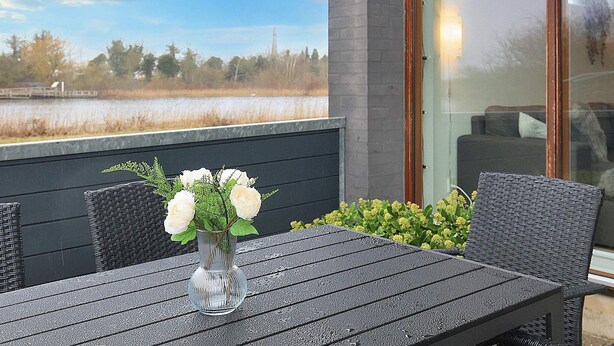 An outdoor patio with a table and chairs, a vase with flowers, and a view of a lake and trees. - 4 Star Holiday Home in Bogense (Bogense)