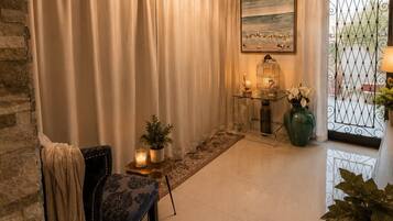 A room with a stone wall, a chandelier, a table with candles, a chair, and a view through a window with a metal gate.