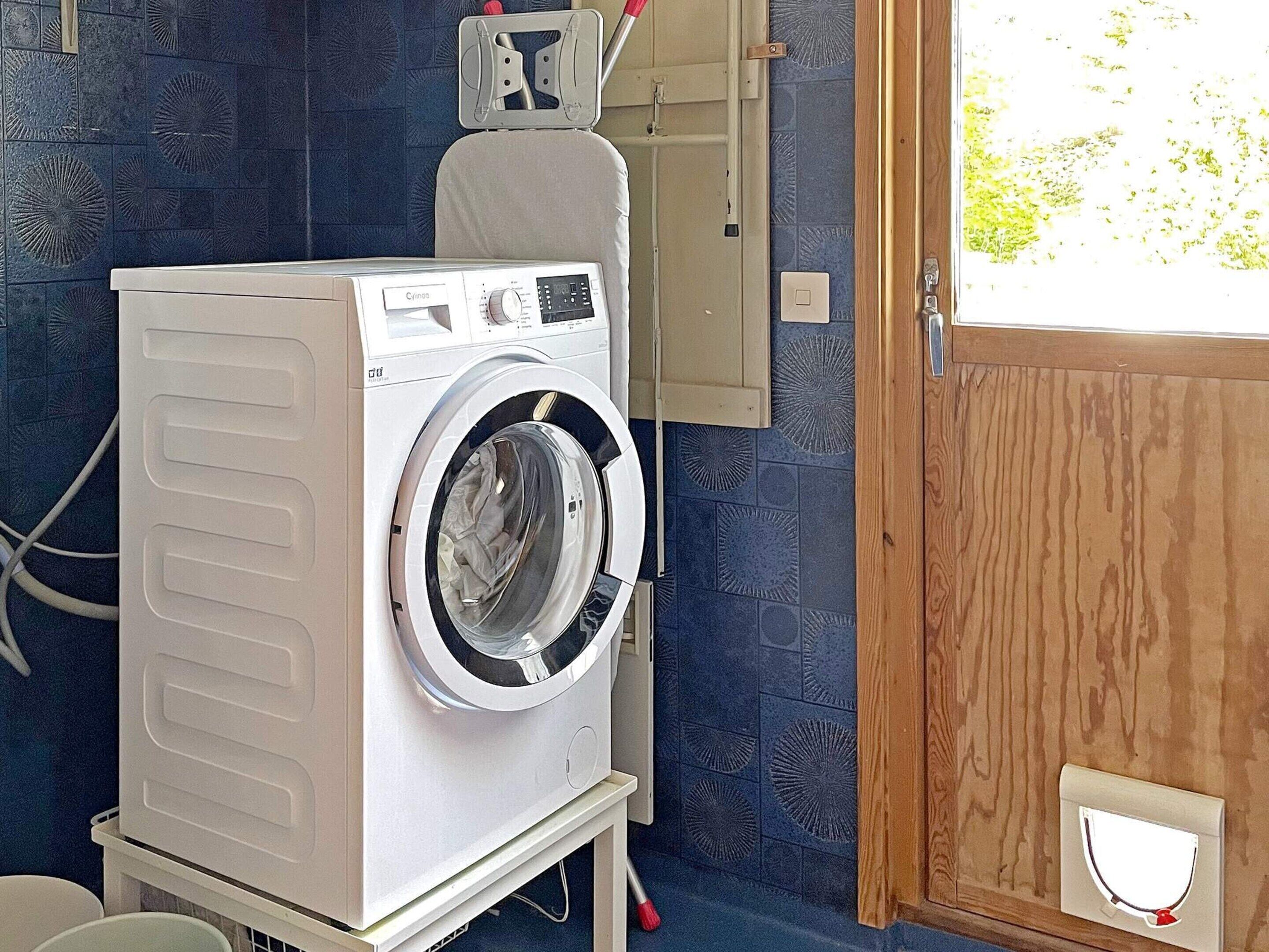 Une petite machine à laver blanche à chargement frontal, installée dans un petit espace buanderie, avec un mur à motifs bleus et une porte en bois.