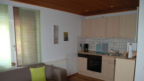 A living room with a sofa and a small table, a kitchen with wooden cabinets and a dishwasher, and a window with blinds.