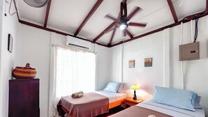 Et hotelværelse med to senge, en loftsvifte, en aircondition, et lille bord med en lampe og en dekorativ skål på et skab.