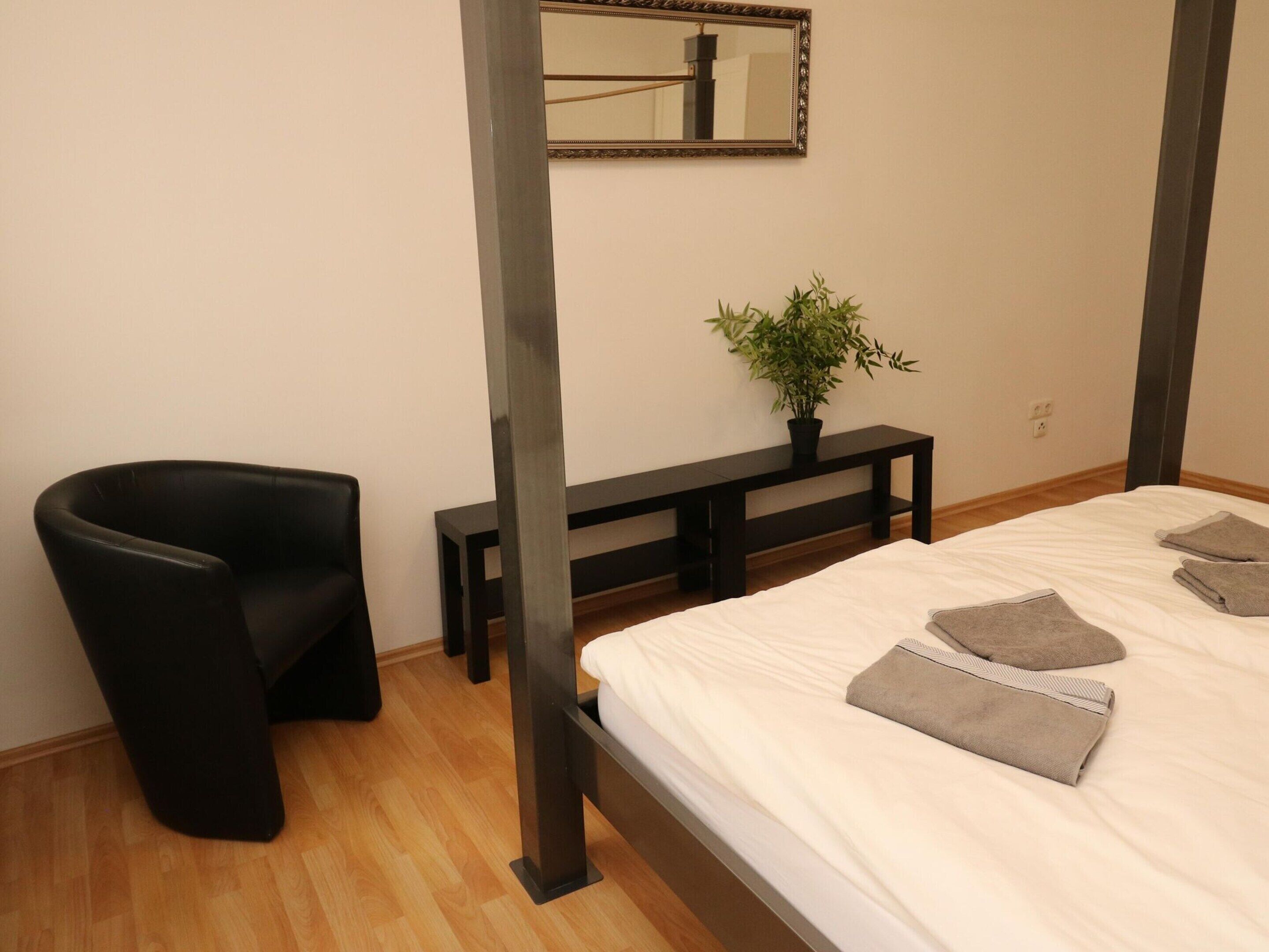 A bedroom with a bed, a black chair, a small table, a potted plant, and a mirror on the wall.